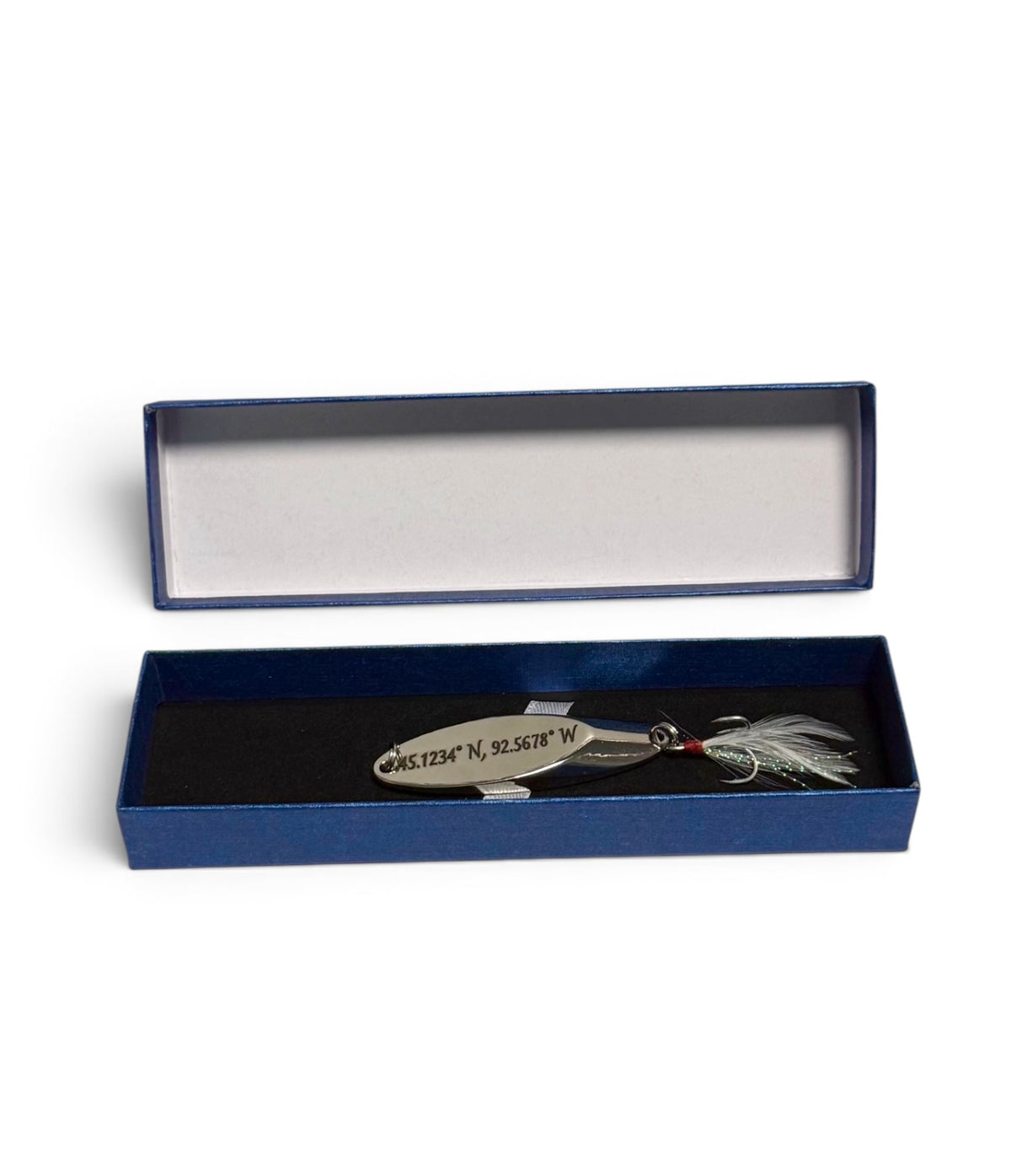 A silver metal fishing lure engraved with geographic coordinates, displayed inside an open dark blue presentation box with a black velvet insert on a white background.