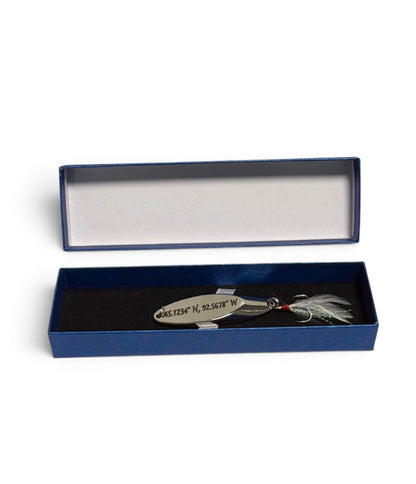 A silver metal fishing lure engraved with geographic coordinates, displayed inside an open dark blue presentation box with a black velvet insert on a white background.