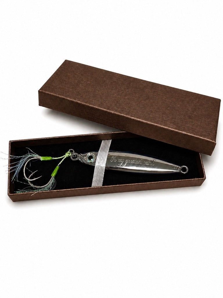 A silver metal fishing lure with a treble hook and green-and-white feather strands, presented in an open brown gift box with the lid leaning behind it on a white background.