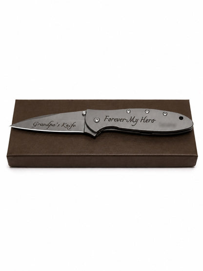 A personalized folding pocket knife with a satin finish, displayed on top of a brown presentation box. The blade is engraved with 'Grandpa's Knife' and the handle with 'Forever My Hero' in script.
