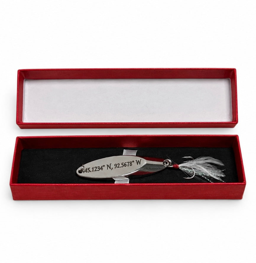 A silver metal fishing lure engraved with geographic coordinates, displayed inside an open red presentation box with a black velvet insert and white lining.