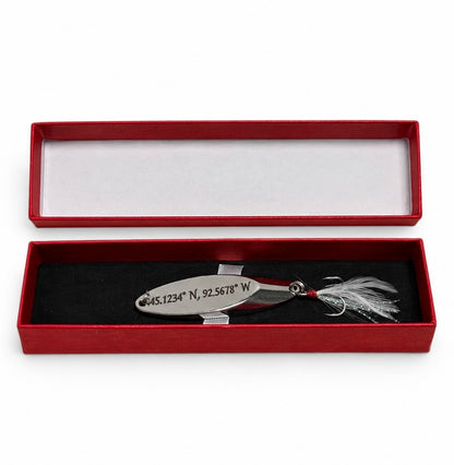 A silver metal fishing lure engraved with geographic coordinates, displayed inside an open red presentation box with a black velvet insert and white lining.