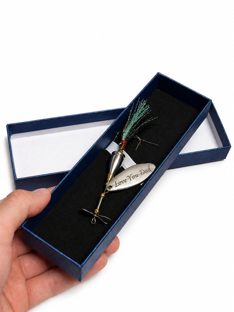 A fishing lure with a silver spinner blade engraved with 'Love You Dad' is presented in an open dark blue gift box with a black velvet insert, held by a hand against a white background.