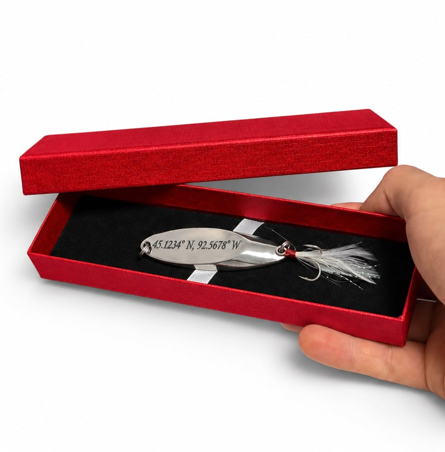 A silver metal fishing lure with engraved coordinates and a white feather jig, presented in an open red gift box held by a hand against a white background.