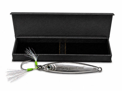 A silver metal fishing lure with a treble hook and white tinsel skirt, displayed in front of an open black presentation box on a white background. The lure has engraved text reading 'For my greatest catch'.