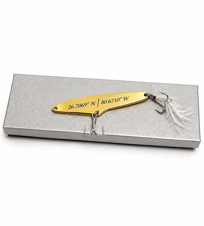 A gold metal spinnerbait fishing lure with engraved coordinates and two treble hooks, displayed on top of a silver rectangular gift box against a white background.