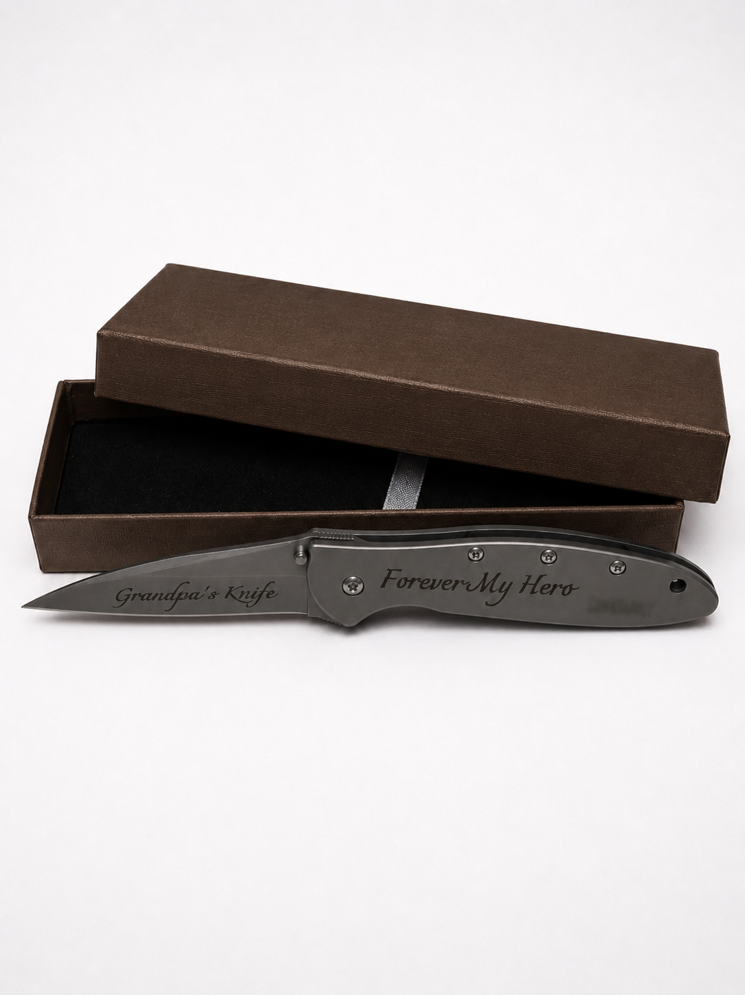 A dark gray personalized folding pocket knife with engraved text 'Grandpa's Knife' and 'Forever My Hero', displayed in front of its brown presentation box on a white background.