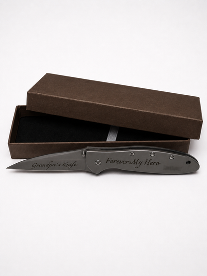 A dark gray personalized folding pocket knife with engraved text 'Grandpa's Knife' and 'Forever My Hero', displayed in front of its brown presentation box on a white background.