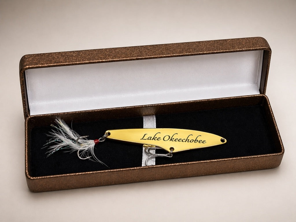 A custom fishing lure engraved with 'Lake Okechobee' rests inside an open brown presentation box with a black velvet lining and white interior.
