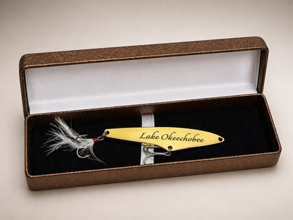 A custom fishing lure engraved with 'Lake Okechobee' rests inside an open brown presentation box with a black velvet lining and white interior.
