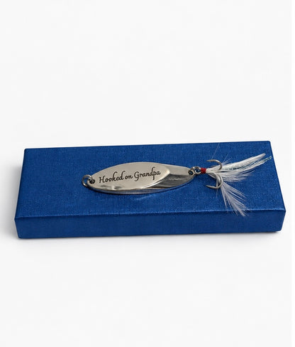A silver metal fishing lure engraved with 'Hooked on Grandpa' and finished with white feathers, displayed on top of a blue rectangular box against a white background.
