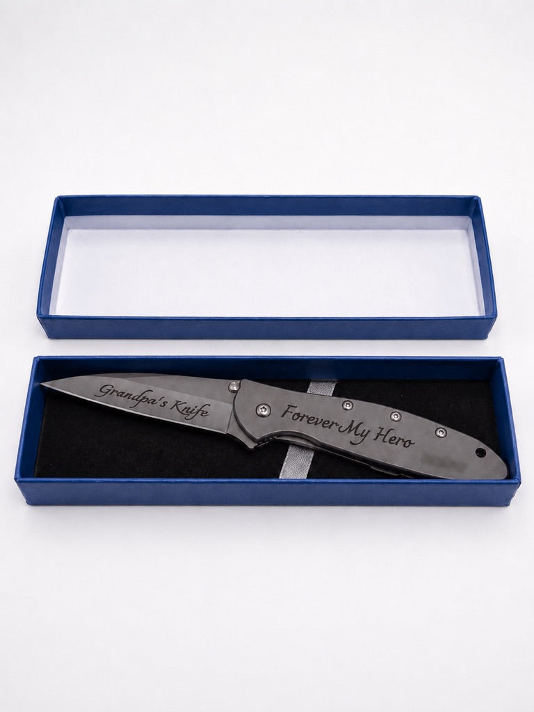 A personalized folding pocket knife with a dark gray metal blade and handle, displayed inside an open blue presentation box with a black velvet insert. The blade is engraved with 'Grandpa's Kaffe' and the handle with 'Forever My Hero'.