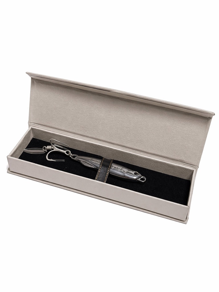 A silver metal fishing lure with a black and silver patterned body, presented inside an open beige gift box with a black insert, on a white background.