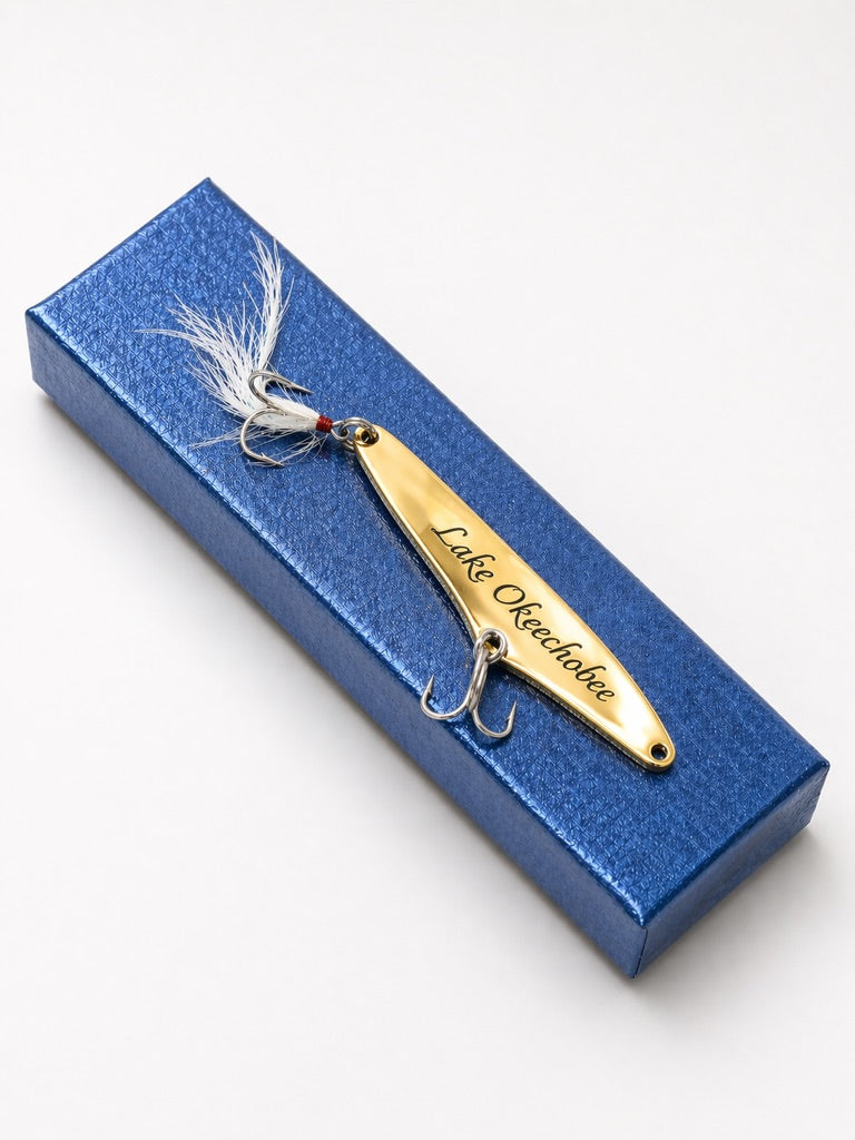 A gold metal fishing lure with white feathered tail and a red bead, engraved with 'Lake Okeechobee', resting on a textured blue gift box against a white background.