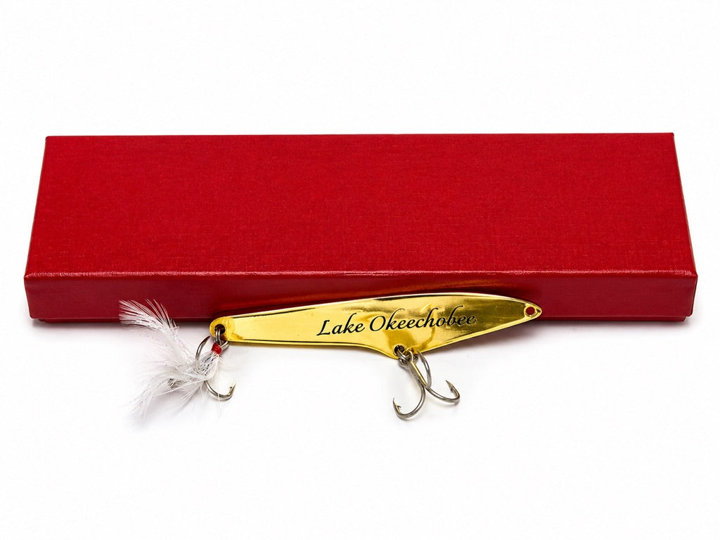 A gold metal fishing lure engraved with 'Lake Okeechobee' and a white feather, displayed in front of a red rectangular presentation box on a white background.