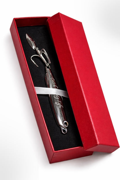 A silver metal fishing lure with integrated hooks, presented inside an open red gift box with a black insert and a white ribbon, on a white background.