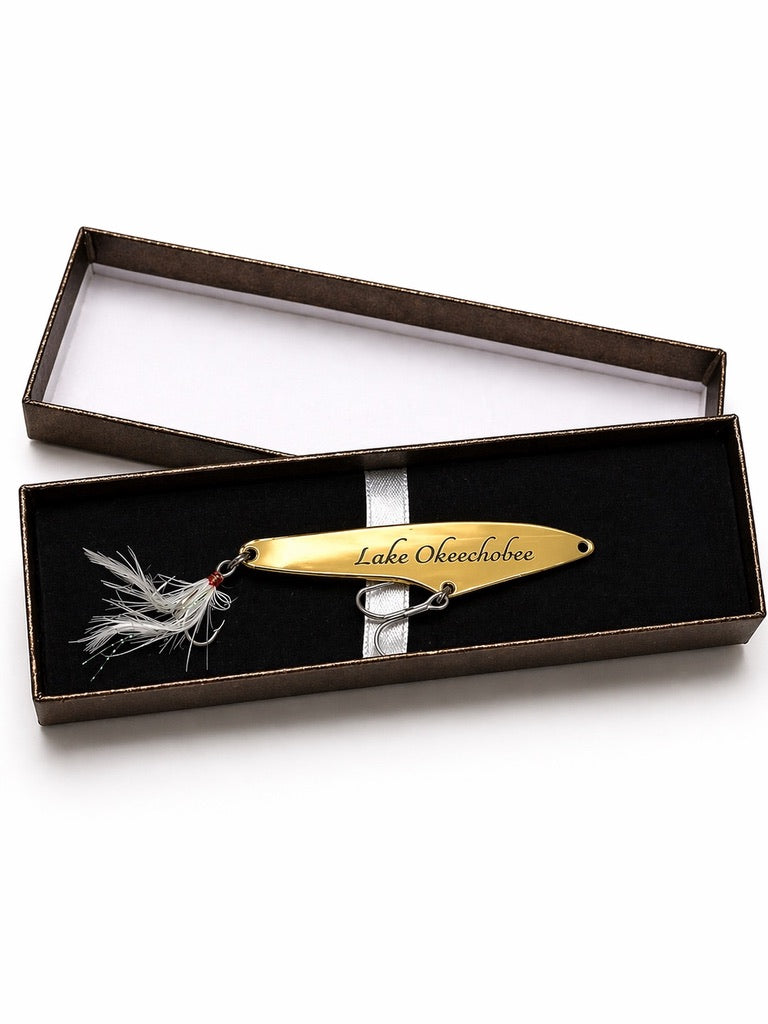 A gold-colored metal fishing lure engraved with 'Lake Ok pochobee' and a white feather jig head, presented in an open dark brown gift box with a black velvet insert on a white background.