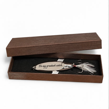 A silver metal fishing lure engraved with 'To my greatest catch.' and displayed inside an open dark brown presentation box with a black insert.