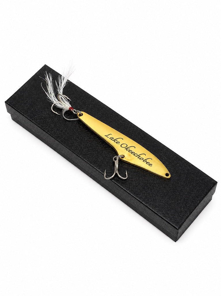 A gold metal fishing lure with white feather accents and two treble hooks, displayed on a black rectangular presentation box. The lure is engraved with the text 'Lake Osculobo'.