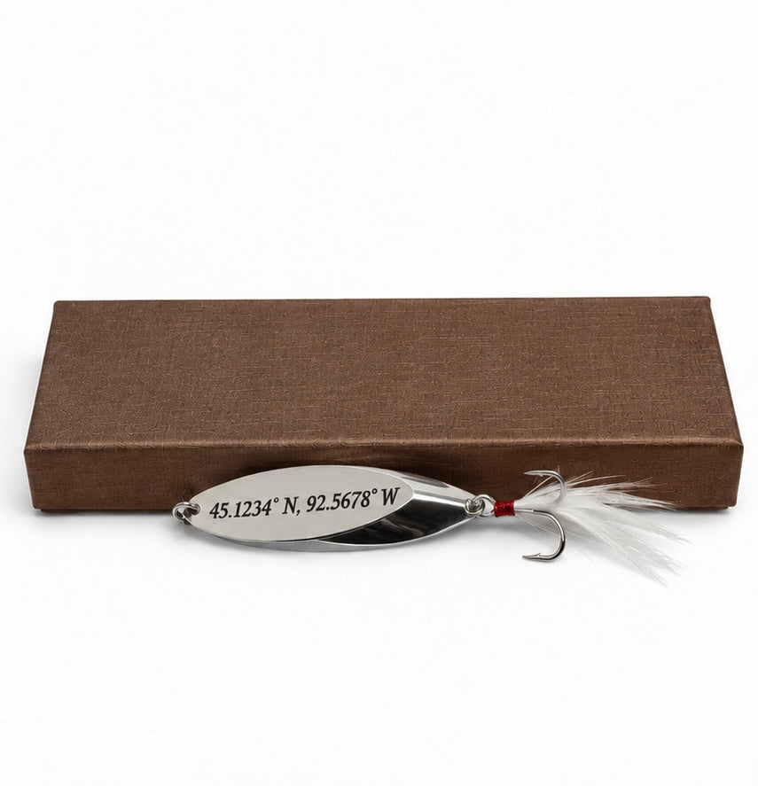 A silver metal fishing lure with white feathers and a treble hook, positioned in front of a brown textured box on a white background. The lure is engraved with geographical coordinates.