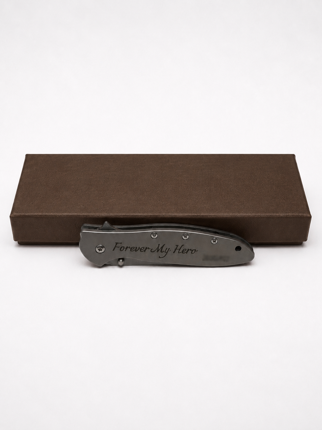 A dark gray folding pocket knife with 'Forever My Hero' engraved on the handle, displayed in front of a brown textured presentation box on a white background.