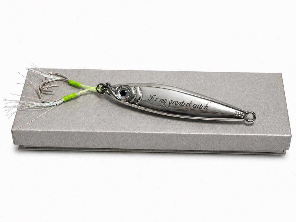 A silver metal fishing lure with a fish-shaped body, engraved with 'For my greatest catch', displayed on a textured gray presentation box against a white background.