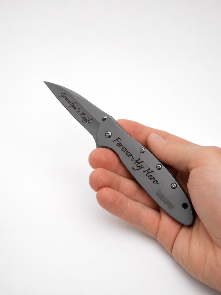 A hand holding a gray folding pocket knife with 'Graves' & 'Kraft' and 'Forever My Hero' engraved on the blade and handle, set against a plain white background.