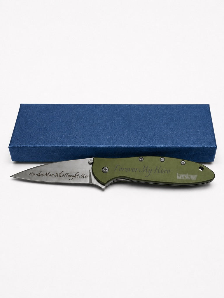 An engraved folding pocket knife with an olive green handle and a silver blade, displayed in front of its textured blue presentation box on a white background.