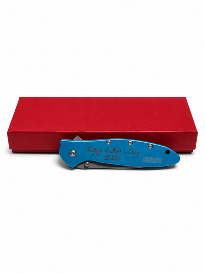 A blue folding pocket knife with 'Happy Father's Day 2026' engraved on the handle, displayed in front of a red presentation box on a white background.