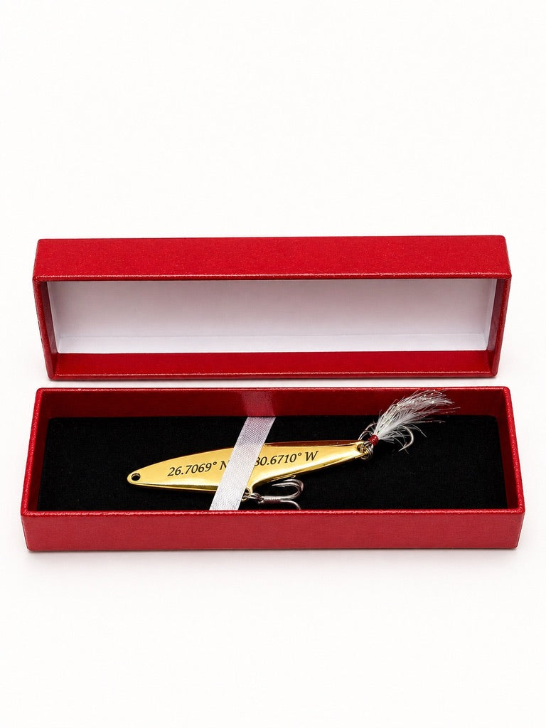 A gold metal fishing lure with a gray feather tail, presented inside an open red gift box with a black velvet insert on a white background.