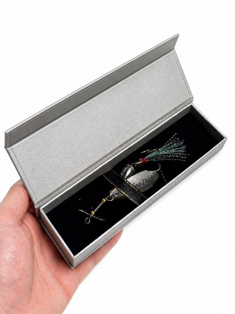 A hand holding an open silver presentation box with a black velvet insert, containing a metallic fishing lure with a black tail and green/red accents.