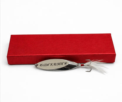A silver metal fishing lure with white feathers and a treble hook, displayed in front of a red textured presentation box on a white background.
