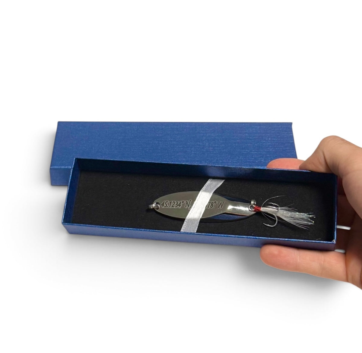 A silver metal fishing lure with a white and red skirt and a white ribbon, presented in an open blue gift box held by a hand against a white background.