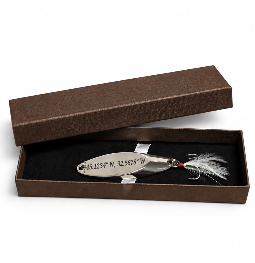 A silver metal fishing spoon lure engraved with geographic coordinates, displayed inside an open brown presentation box with a black insert.