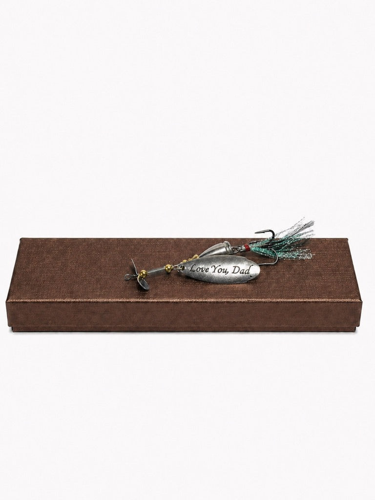 A silver metal fishing lure engraved with 'I Love You Dad' in black script, featuring a green and black feather skirt and a small red bead, displayed on top of a brown textured rectangular gift box against a white background.