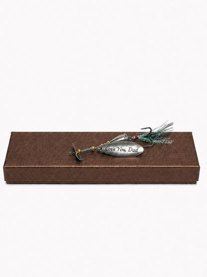 A silver metal fishing lure engraved with 'I Love You Dad' in black script, featuring a green and black feather skirt and a small red bead, displayed on top of a brown textured rectangular gift box against a white background.