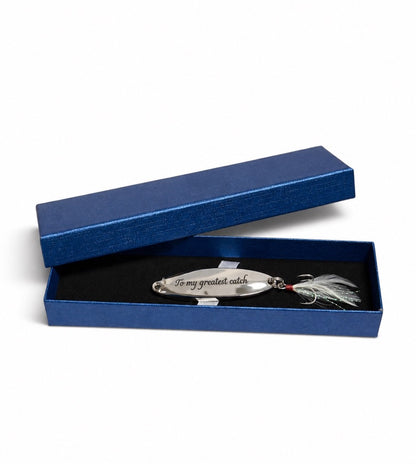 A silver metal fishing lure engraved with 'To my greatest catch' resting inside an open textured blue presentation box with a black velvet lining.
