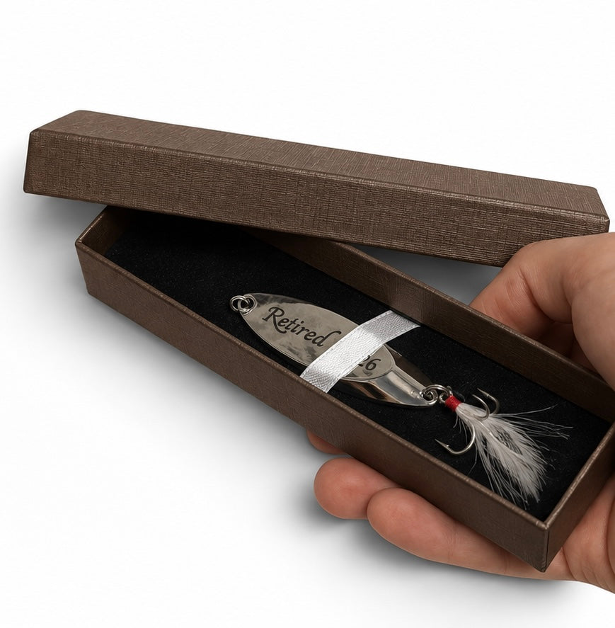 A silver engraved fishing lure with white feathers and a red eye, presented in an open brown gift box held by a hand. The box lid is engraved with 'Retired Cat 16'.