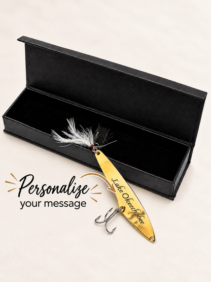 A gold metal fishing lure with white and black feather accents and two silver hooks, displayed in front of an open black presentation box on a light beige background. Text on the image reads 'Personalize your message' and the lure is engraved with 'Lake Overlubber'.