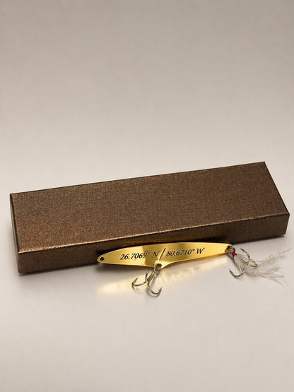 A gold metal fishing lure with two treble hooks and white feather tails, displayed in front of its textured brown presentation box on a neutral light gray background.