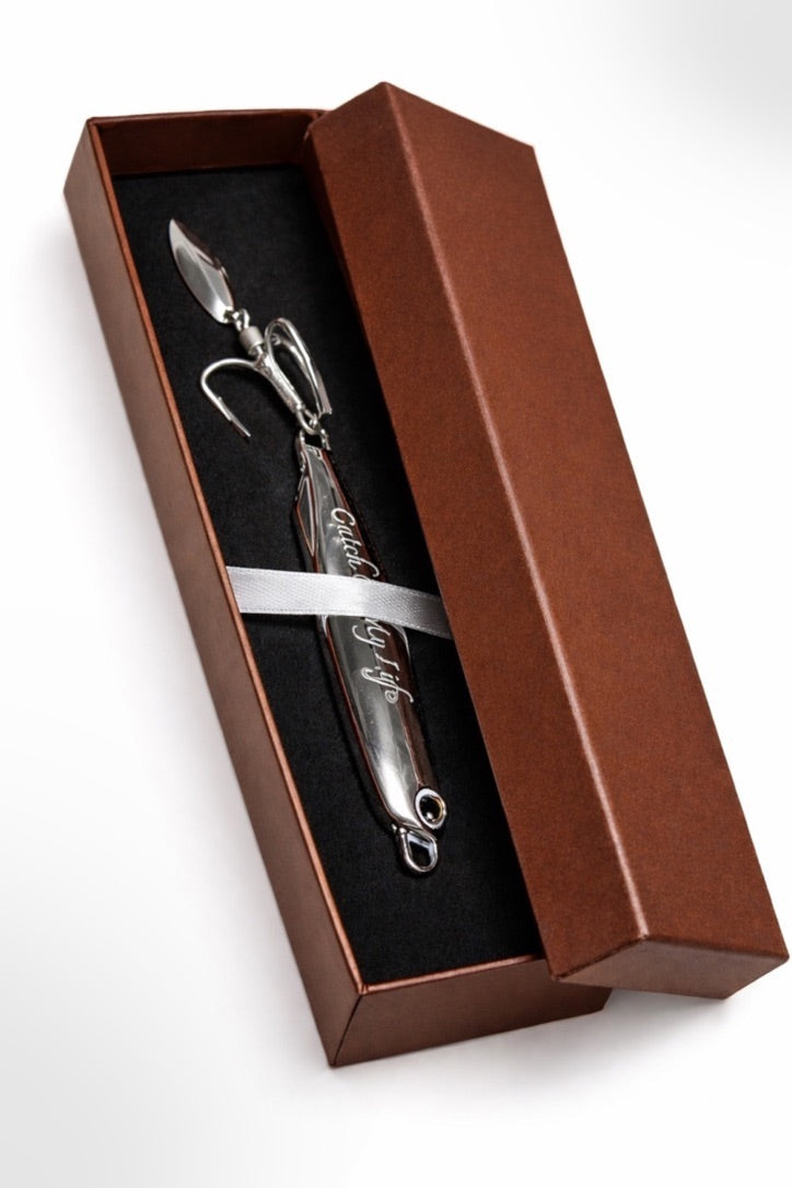 A silver metal fishing lure presented inside an open brown gift box with a black insert and a white ribbon, photographed on a clean white background.