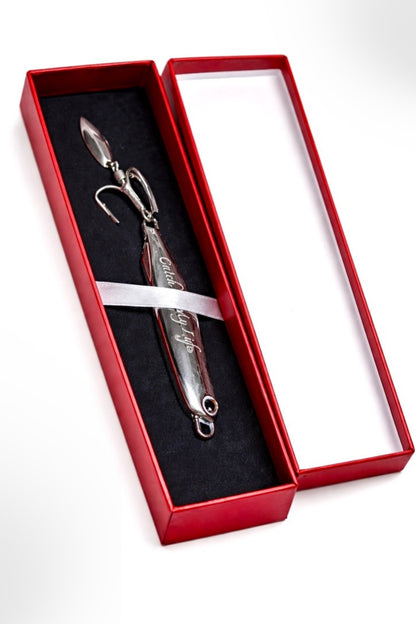 A silver metal fishing lure with engraved script, presented inside an open red gift box with a black velvet insert and a white ribbon, on a white background.