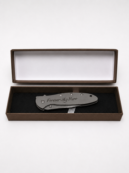 A folding pocket knife with 'Forever My Hero' engraved on the handle, presented inside an open brown gift box with a black velvet insert on a white background.