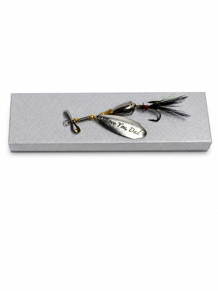 A fishing lure with a silver blade engraved with 'Believe You Can' and a black feathered tail, displayed on a textured gray rectangular box against a white background.