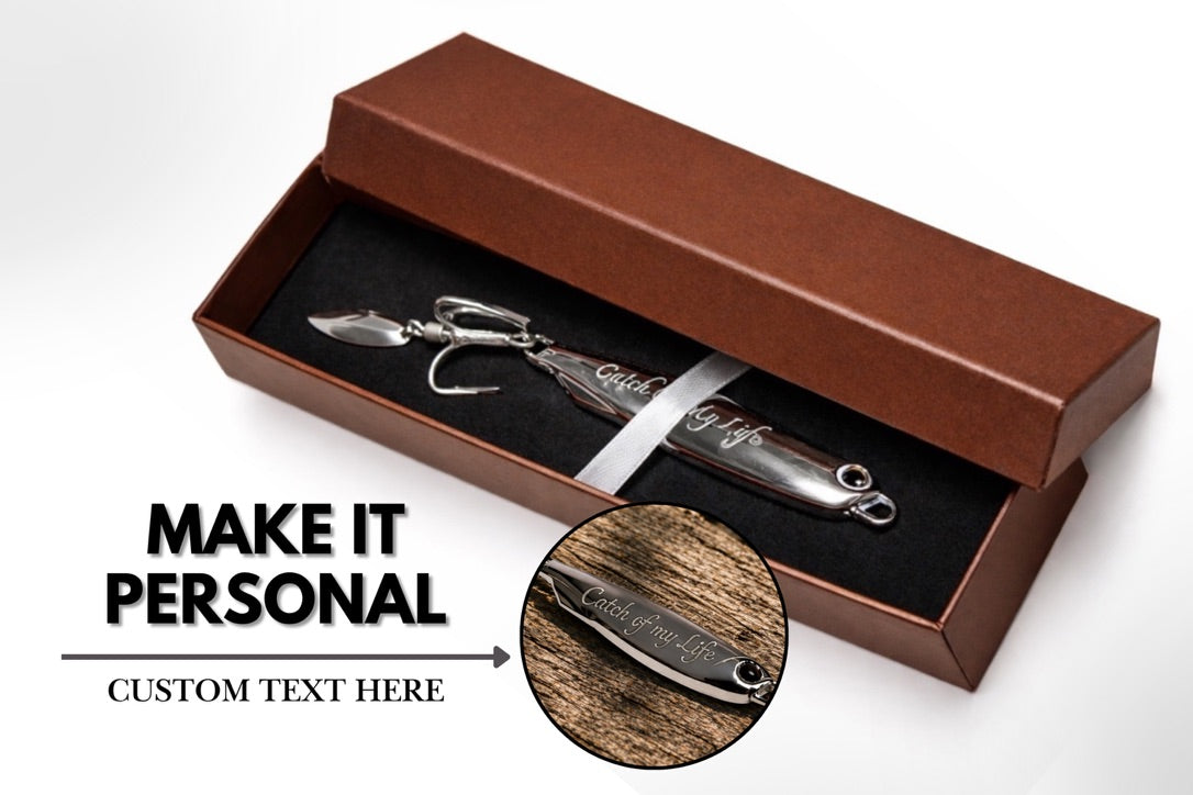 A silver metal fishing lure displayed inside an open brown presentation box with a black insert. The image includes promotional text reading 'MAKE IT PERSONAL' and a circular inset showing engraved wood sample text area, on a clean white background.