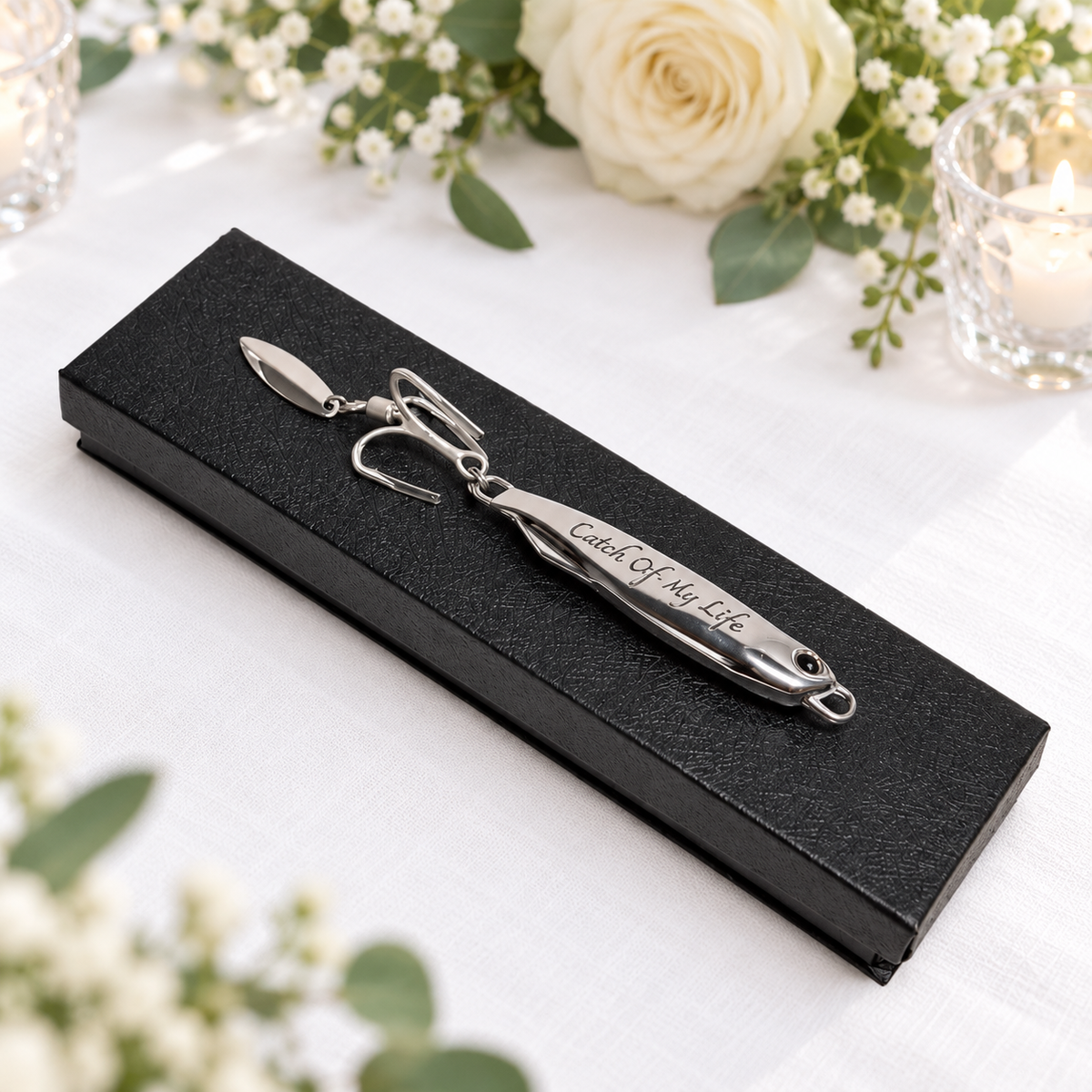 A silver metal fishhook-style keepsake pin engraved with 'Catch Of My Life' rests on a black textured presentation box, styled on a white fabric surface with flowers and candles in the background.