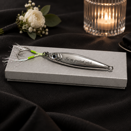 A silver metal fishing lure engraved with 'For my greatest catch' rests on a textured gray gift box, set against a dark draped fabric background with a lit candle and small white flowers in the background.