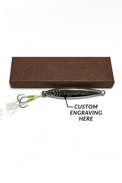 A silver metal fishing lure with green and white skirt strands, shown in front of a brown rectangular box on a white background. A black text label reads 'CUSTOM ENGRAVING HERE' with an arrow pointing to the lure's body.