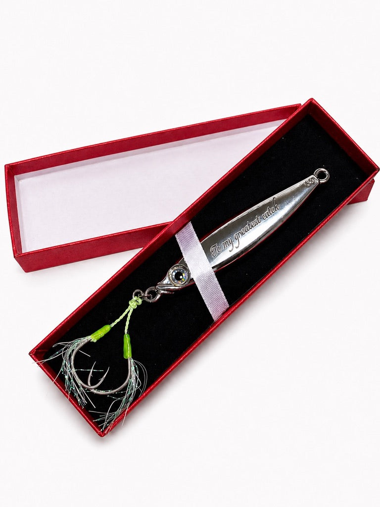 A silver metal fishing lure with green and white feathered skirt and a small blue eye detail, presented in an open red gift box with a black velvet insert.