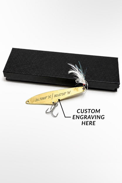 A gold metal fishing lure with a white feather and two hooks, displayed next to a black textured box on a white background. A text overlay reads 'CUSTOM ENGRAVING HERE' with an arrow pointing to the engraved text on the lure.